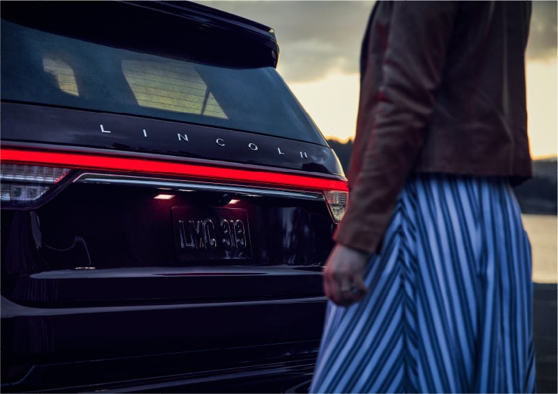 A person is shown near the rear of a 2023 Lincoln Aviator® SUV as the Lincoln Embrace illuminates the rear lights | Gray-Daniels Lincoln in Brandon MS