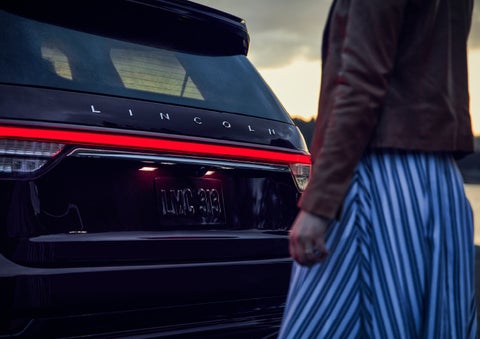 A person is shown near the rear of a 2024 Lincoln Aviator® SUV as the Lincoln Embrace illuminates the rear lights | Gray-Daniels Lincoln in Brandon MS